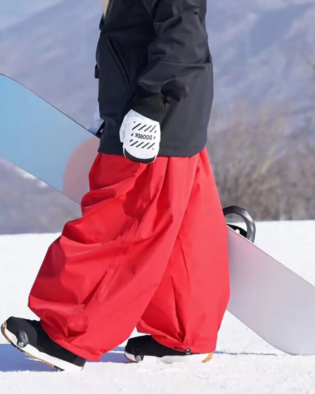 Snowboarder in red baggy ski trousers carrying a snowboard with a scenic mountain backdrop