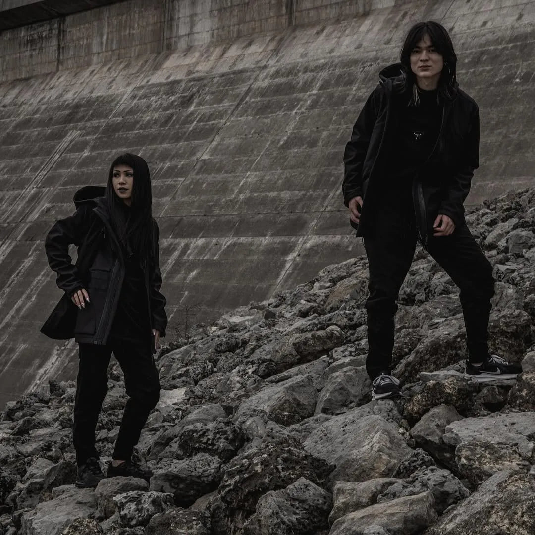 Two models wearing all-black techwear outfits standing on rocks in an outdoor urban landscape, Imaphotic community style feature.