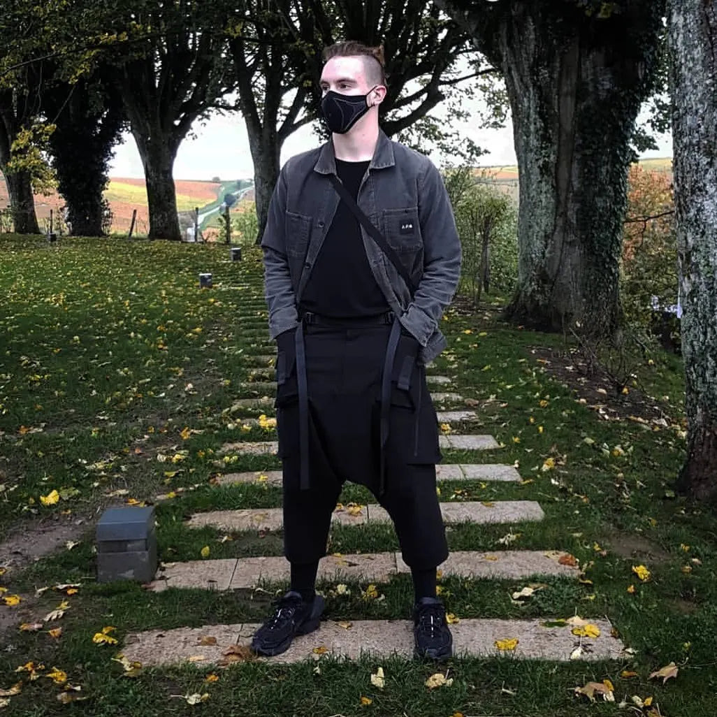 Male model wearing a techwear mask, layered black outfit, and tactical pants in a scenic park with autumn trees – Imaphotic street style.