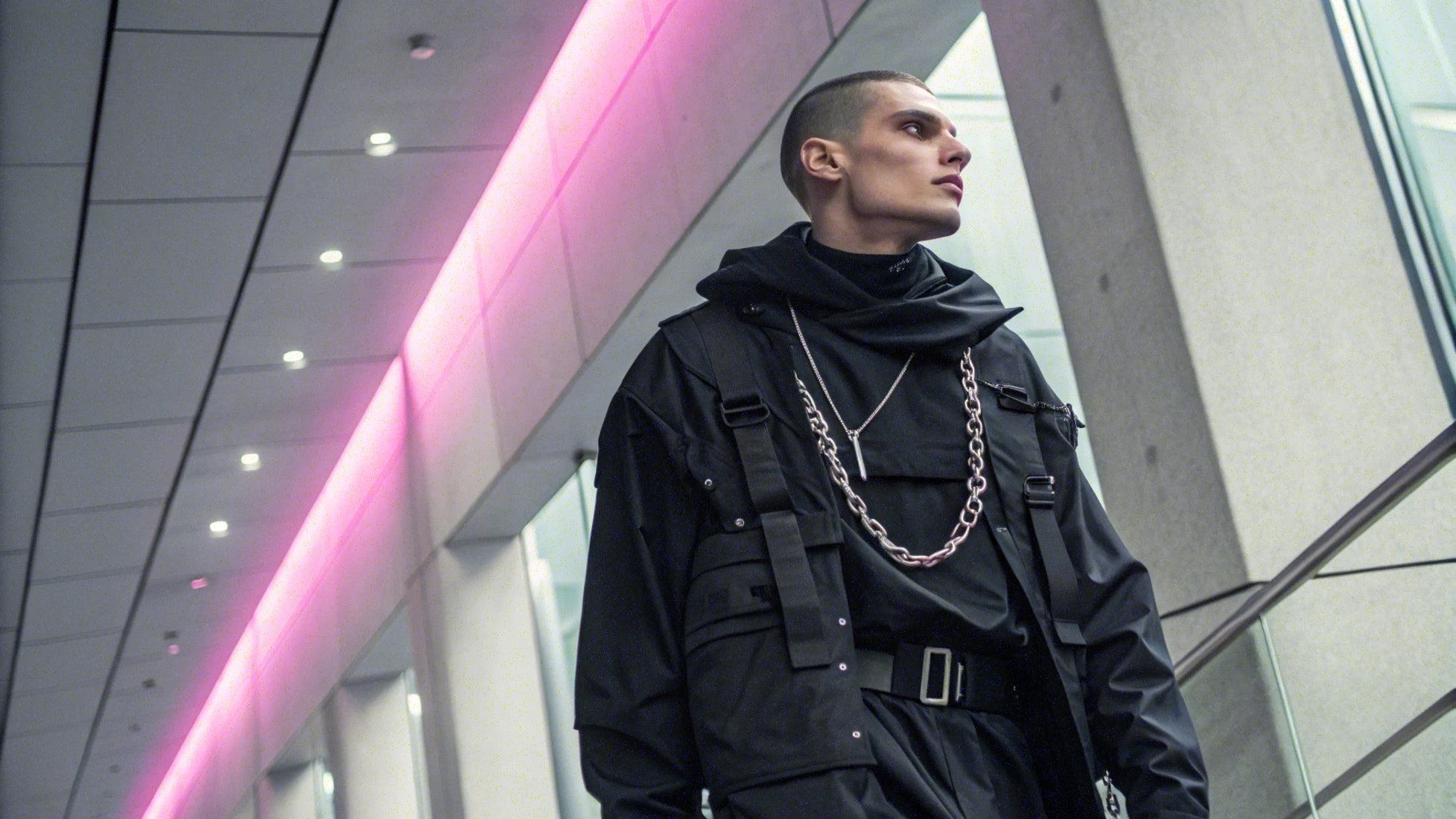 Male model wearing futuristic black techwear outfit with chain accessories, standing under pink neon lights – Imaphotic fashion banner