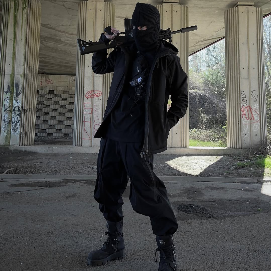 Model in tactical boots and techwear gear holding a prop rifle, styled in an urban underpass with graffiti, Imaphotic underground fashion.